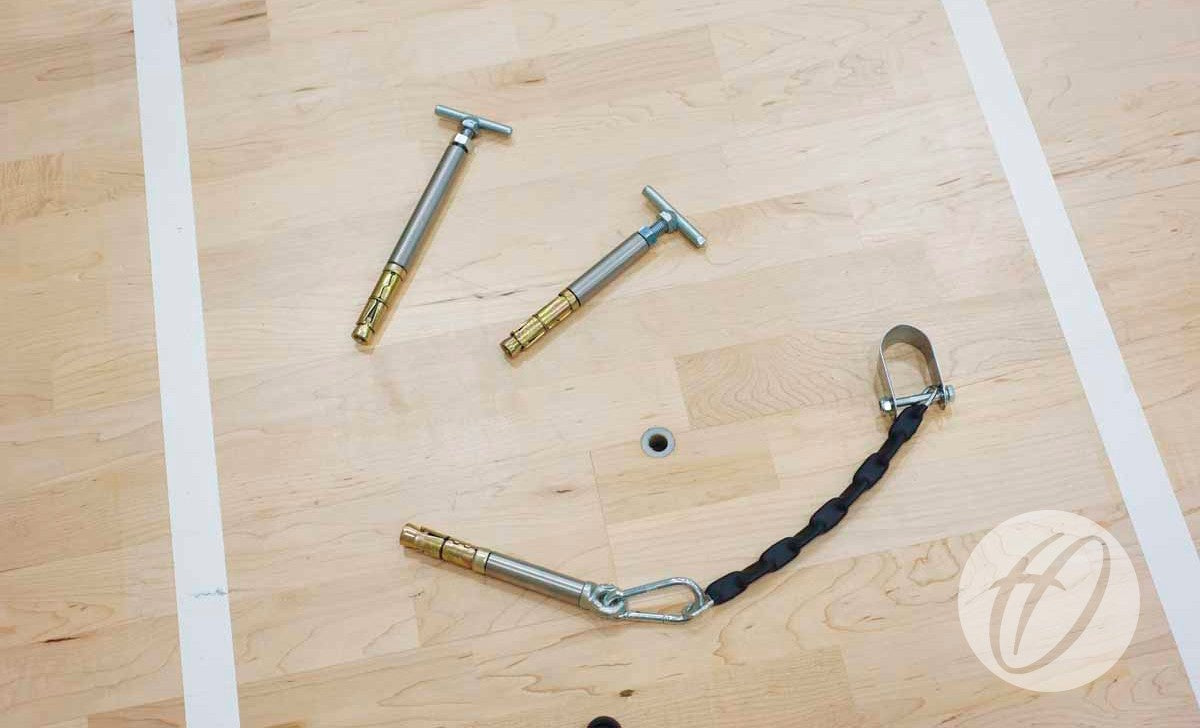 Three Harrod Sport SH2 Sportshall Anchors for suspended floors (210MM) lie on a light wooden gym floor—two loose, one attached to a black strap. White court lines and a circular logo are visible on the right side.
