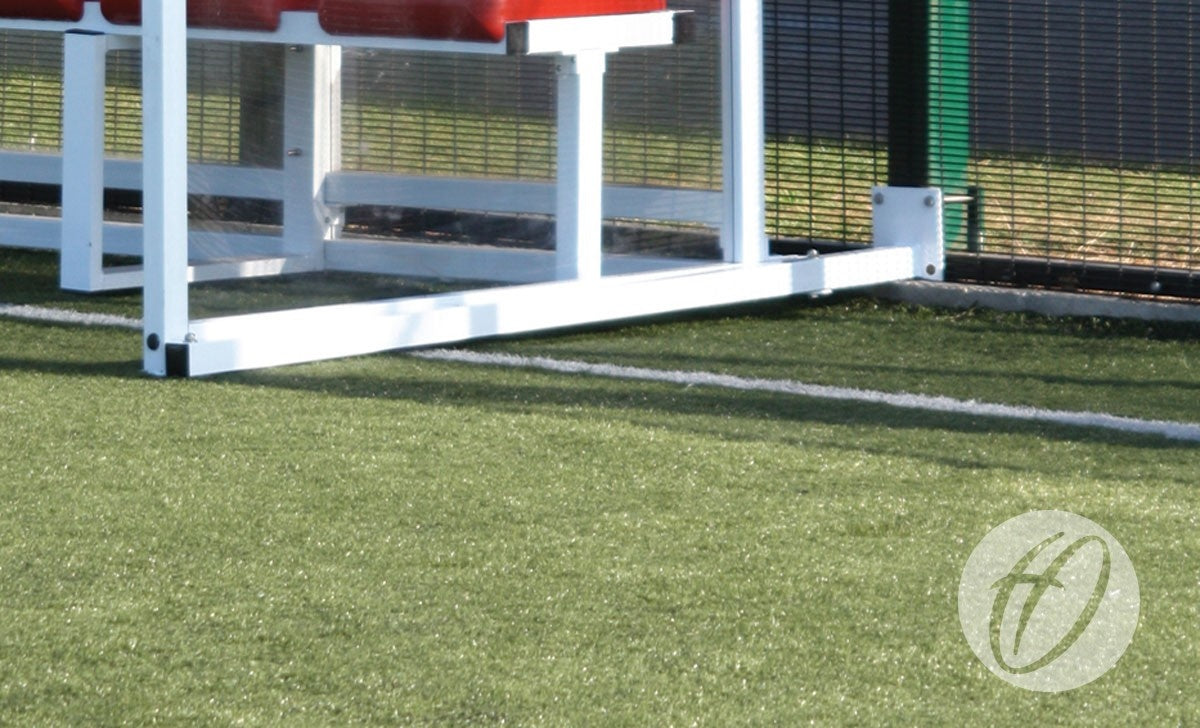 A close-up of the lower section of a white metal bench with red seats on artificial turf by a black wire fence, featuring Harrod Sport Anchor For Standard Shelters near the logo in the bottom right corner.