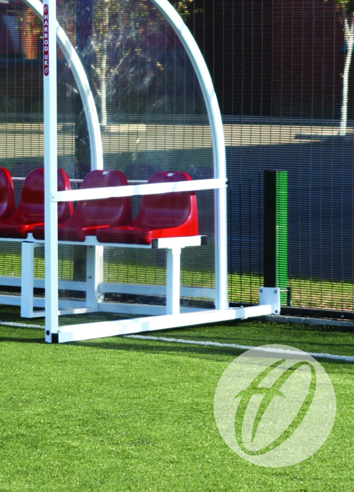 A Harrod Sport Anchor for Premier Shelters secures red plastic seats beneath a white shelter with galvanized steel posts on the edge of artificial green turf, beside a black fence and tall green post.