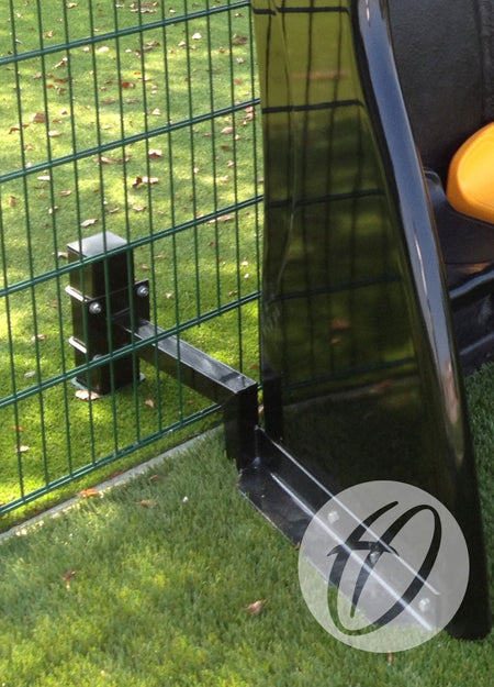 A close-up of a Harrod Sport Fibretech Shelter Post Anchor securing a metal fence panel to artificial grass with galvanised steel posts. A black and yellow item is visible on the right, with brown leaves scattered on the grass.