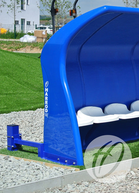 A Harrod Sport Fibretech Shelter Post Anchor with white seats stands on artificial grass beside gravel, supported by galvanised steel posts. A building and trees can be seen in the background.