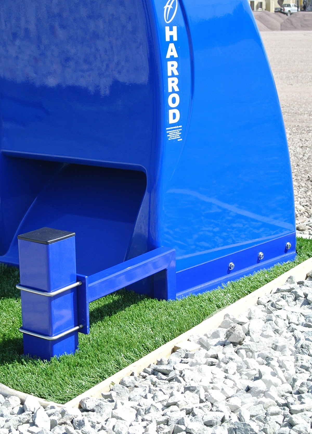 A close-up of a bright blue Harrod Sport Fibretech Shelter Post Anchor with galvanized steel posts on artificial grass, bordered by white edging and gray stones.
