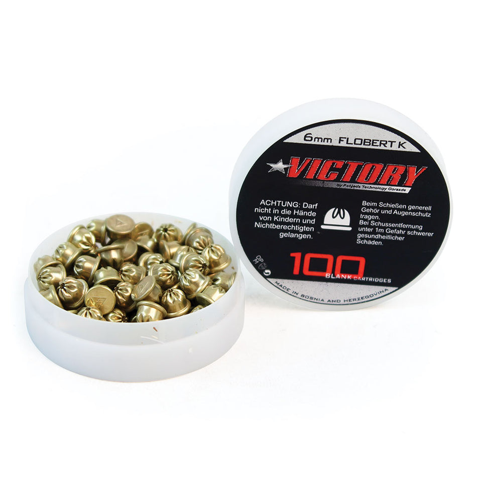 A round white container of Martin Berrill Sports Starting Pistol Blanks is shown open, revealing brass cartridges. The lid displays a black-and-red label with “6mm Flobert K VICTORY” and warning text in German.