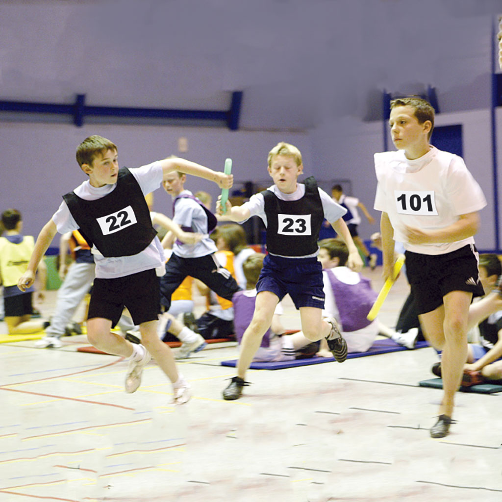 Three boys wearing numbered bibs run indoors in a relay race. One passes a green Mastersport Plastic Relay Baton (from the Set of Six) to a teammate, while another runs ahead. Spectators and participants watch from the background.