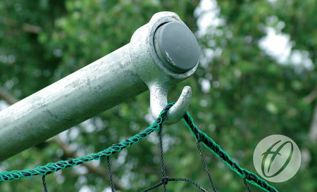 Close-up of a metal hook securing a Harrod Sport Practice Discus Cage Net with green and black square mesh to a horizontal pole. Blurred green foliage is visible in the background, with the semi-transparent circular logo at the bottom right.