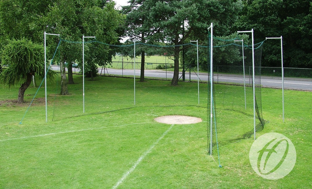 The Harrod Sport Practice Discus Cage Net, featuring square mesh polypropylene and metal poles, encloses a concrete throwing circle on grass, with trees and a road visible in the background.