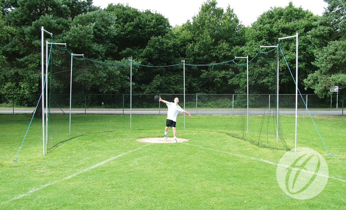 Wearing athletic gear, a person prepares to throw a discus inside the Harrod Sport Practice Discus Cage Net, made of square mesh polypropylene, set on a grassy field with trees and a fence under sunny skies.