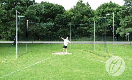 Wearing athletic gear, a person prepares to throw a discus inside the Harrod Sport Practice Discus Cage Net, made of square mesh polypropylene, set on a grassy field with trees and a fence under sunny skies.
