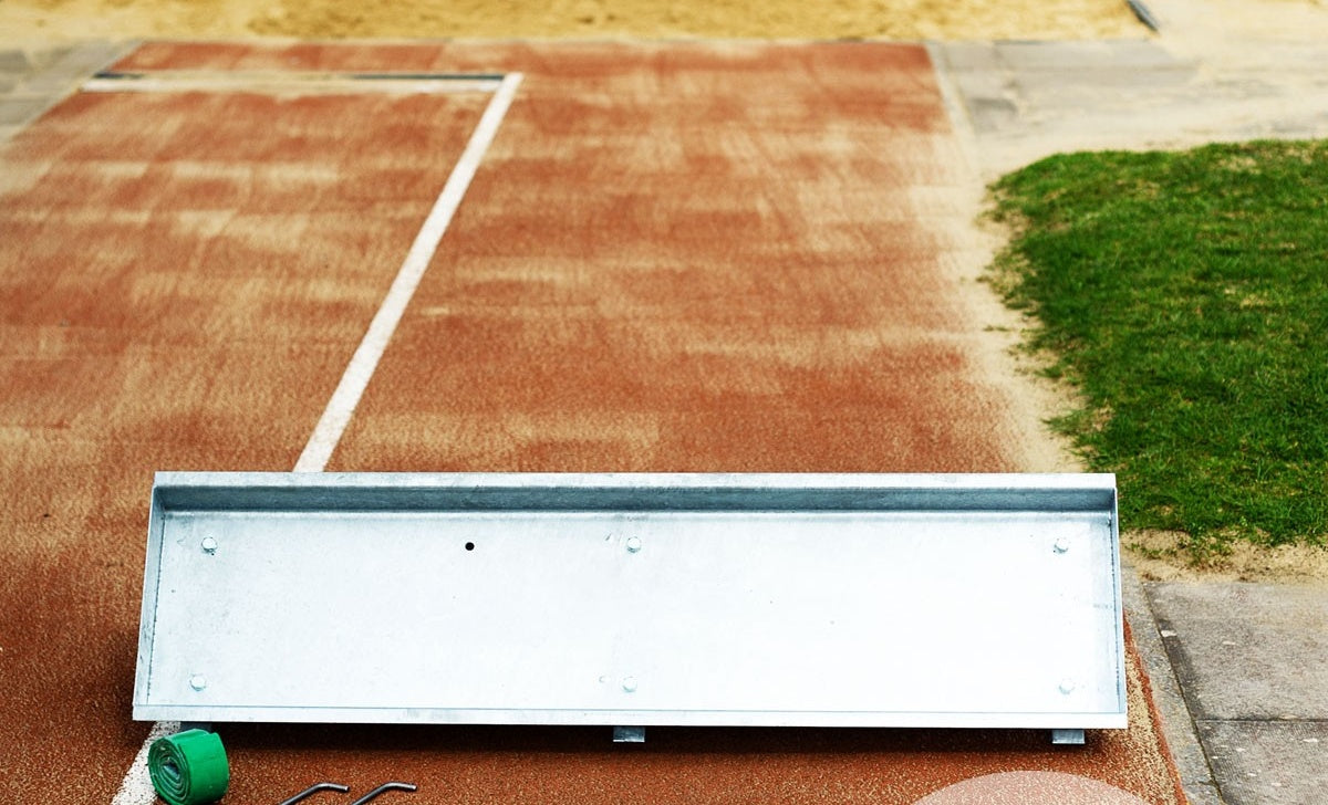 A Harrod Sport Long Jump Trough (1210mm, galvanised steel) is installed in the sandpit. In the foreground are a metal barrier and a green rolled-up mat. Grass borders the runway on the right.