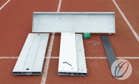 Harrod Sport Blanking Board metal components and tools for athletic track assembly, including long jump foundations, are arranged on a red running track with white lane markings. Also shown: a green measuring tape and hex keys.