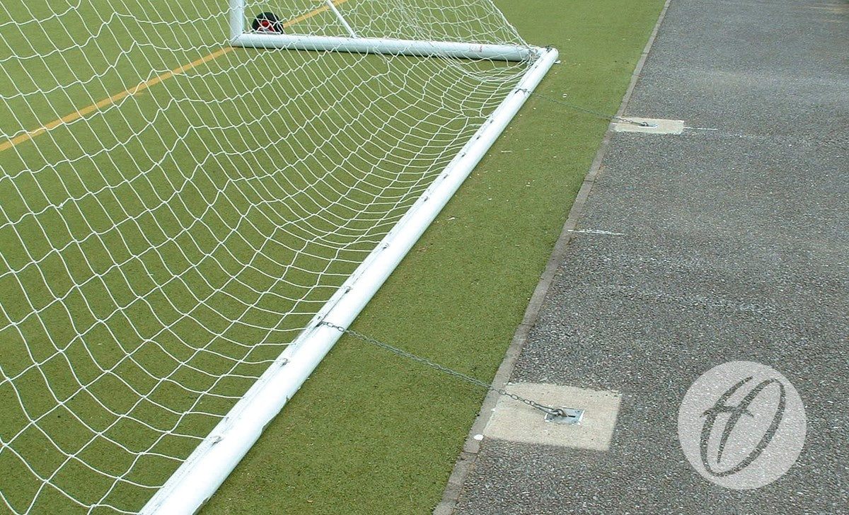 A Harrod Sport Fence Anchor Chain secures a soccer goalpost with carbine clips on artificial turf near pavement. The curved H logo appears in the lower right corner.