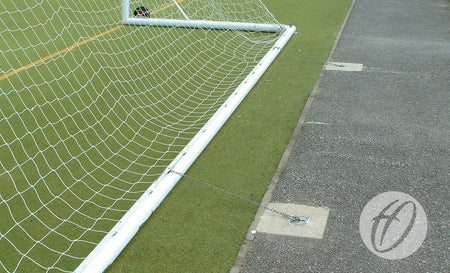 A Harrod Sport Fence Anchor Chain secures a soccer goalpost with carbine clips on artificial turf near pavement. The curved H logo appears in the lower right corner.