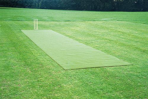 The Huck Nets Refurbishment of Artificial Wicket features a green mat cricket pitch with wooden stumps, set in a grassy field surrounded by trees—ideal for upgrading or maintaining artificial wickets.