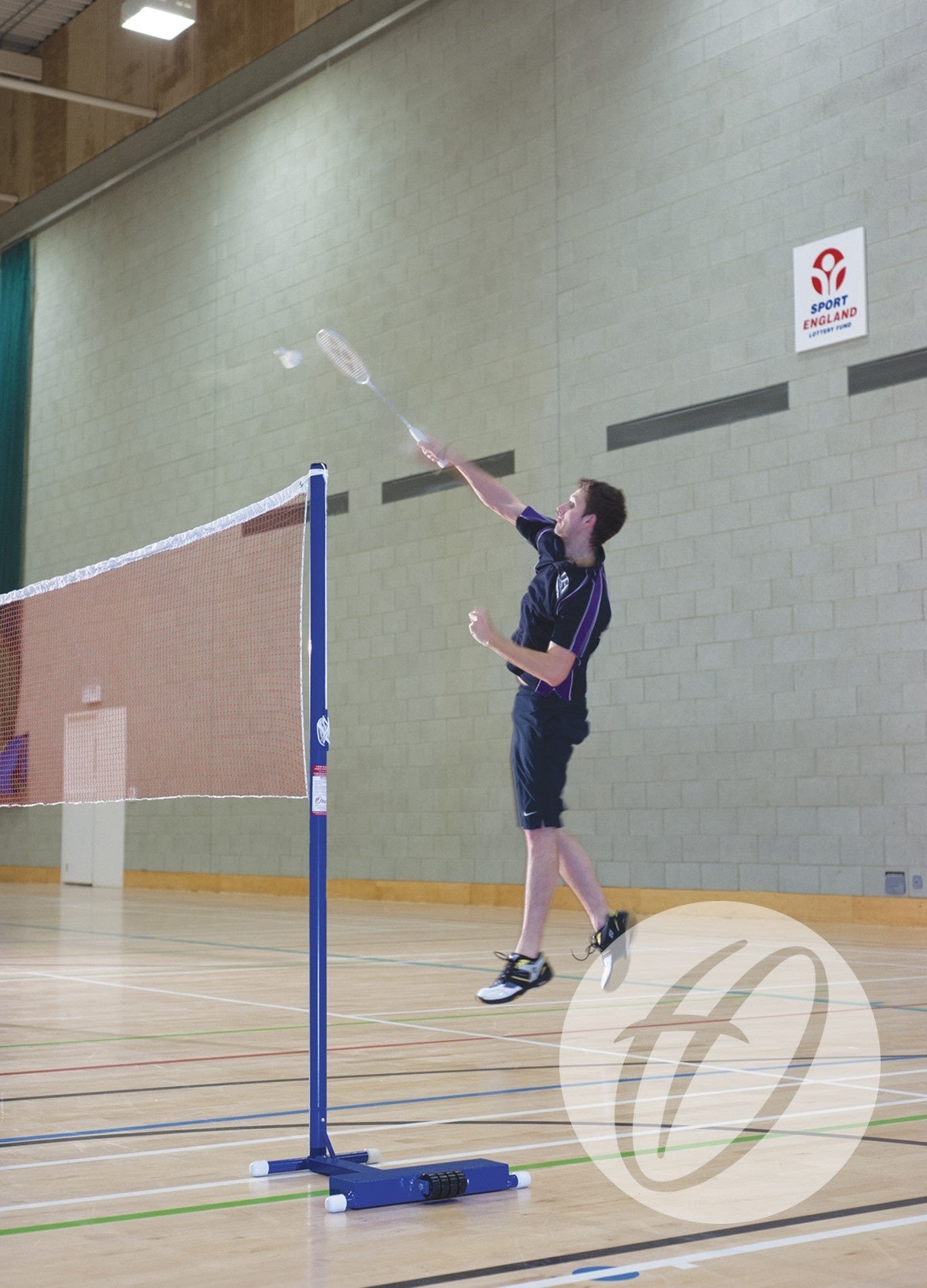 A man in sportswear jumps to hit a shuttlecock with a badminton racket on an indoor court. Harrod Sport Club Wheelaway Training Posts and a net are visible, with a Sport England sign suggesting club use of the facility.