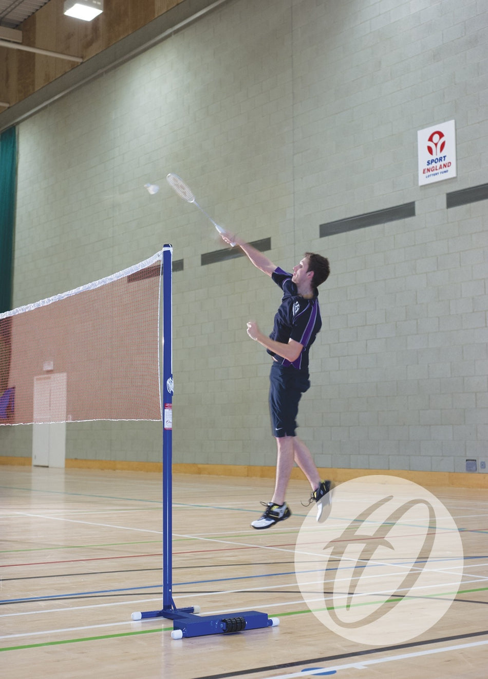 A man in sportswear jumps to hit a shuttlecock with a badminton racket on an indoor court. Harrod Sport Club Wheelaway Training Posts and a net are visible, with a Sport England sign suggesting club use of the facility.