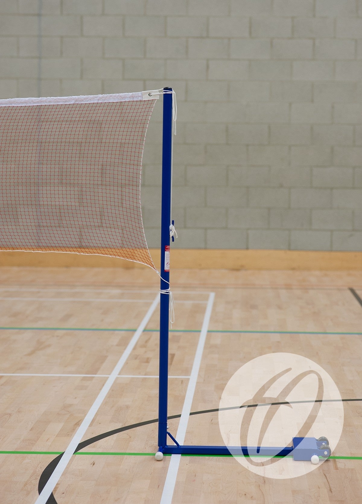 The Harrod Sport School Wheelaway Training Posts, a blue portable badminton net system, stand on a wooden gym floor with court lines, ideal for clubs and schools, set against a light gray brick wall.