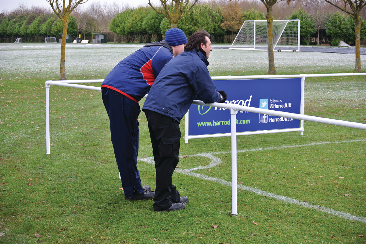 Two people in winter clothing lean on a Harrod Sport 2.1M Spectator Barrier Mid-section, watching a sports field with goalposts and frosty grass. A blue banner is attached to the fence, and trees line the background.