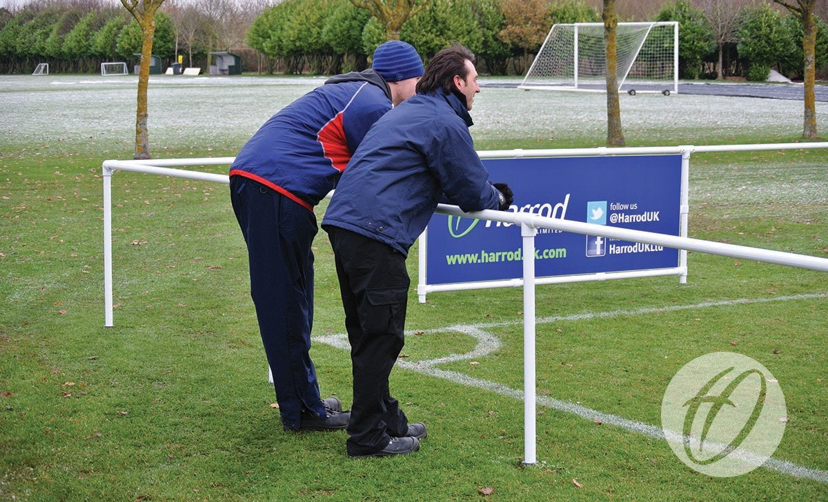Two people in winter clothing lean on a Harrod Sport 2.1M Spectator Barrier Corner, watching a grassy soccer field. A blue advertisement banner is attached to the barrier on this chilly day.