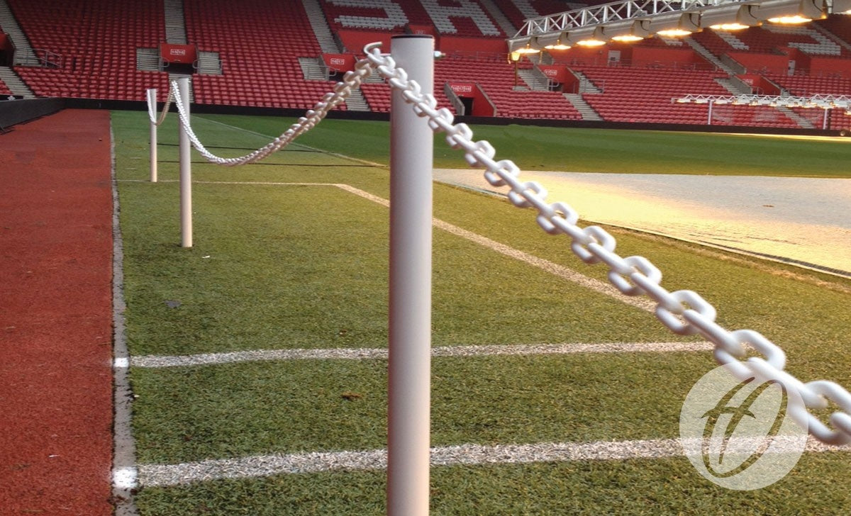 A Harrod Sport Pitch Protection Barrier, featuring a white plastic chain on posts, separates the soccer field from the stands in a mostly empty stadium with red seats and artificial turf visible—an installation reminiscent of Harrod Sport products.
