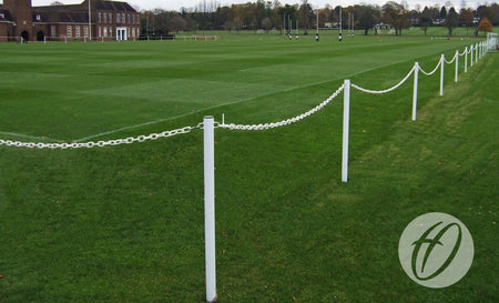 A green sports field featuring a Harrod Sport Pitch Protection Barrier with a white plastic chain, a brick building in the background, and distant trees. A few people are visible near the far end of the field, possibly using additional equipment from Harrod Sport.
