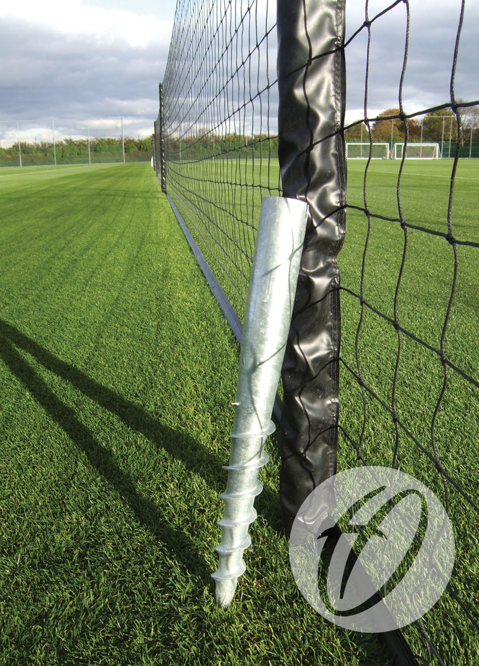 A Harrod Sport Turf Screw Socket & Cap is installed at the base of a net post on a grass soccer field, securing the net. The field is marked for soccer, and goals can be seen in the background.