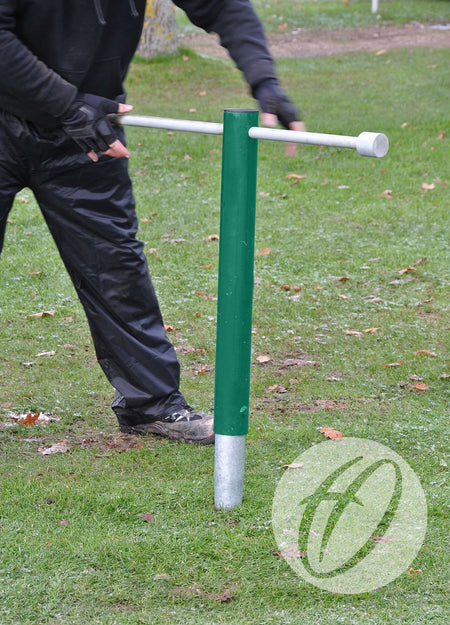 Wearing black gloves and pants, a person uses the Harrod Sport Turf Screw Insertion Tool to install or remove a green metal post anchored in grassy ground with scattered leaves.