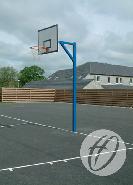 A Harrod Sport Heavy Duty Basketball Goal with a steel backboard and blue pole stands on an asphalt outdoor court; a wooden fence is visible in the background beneath a cloudy sky.