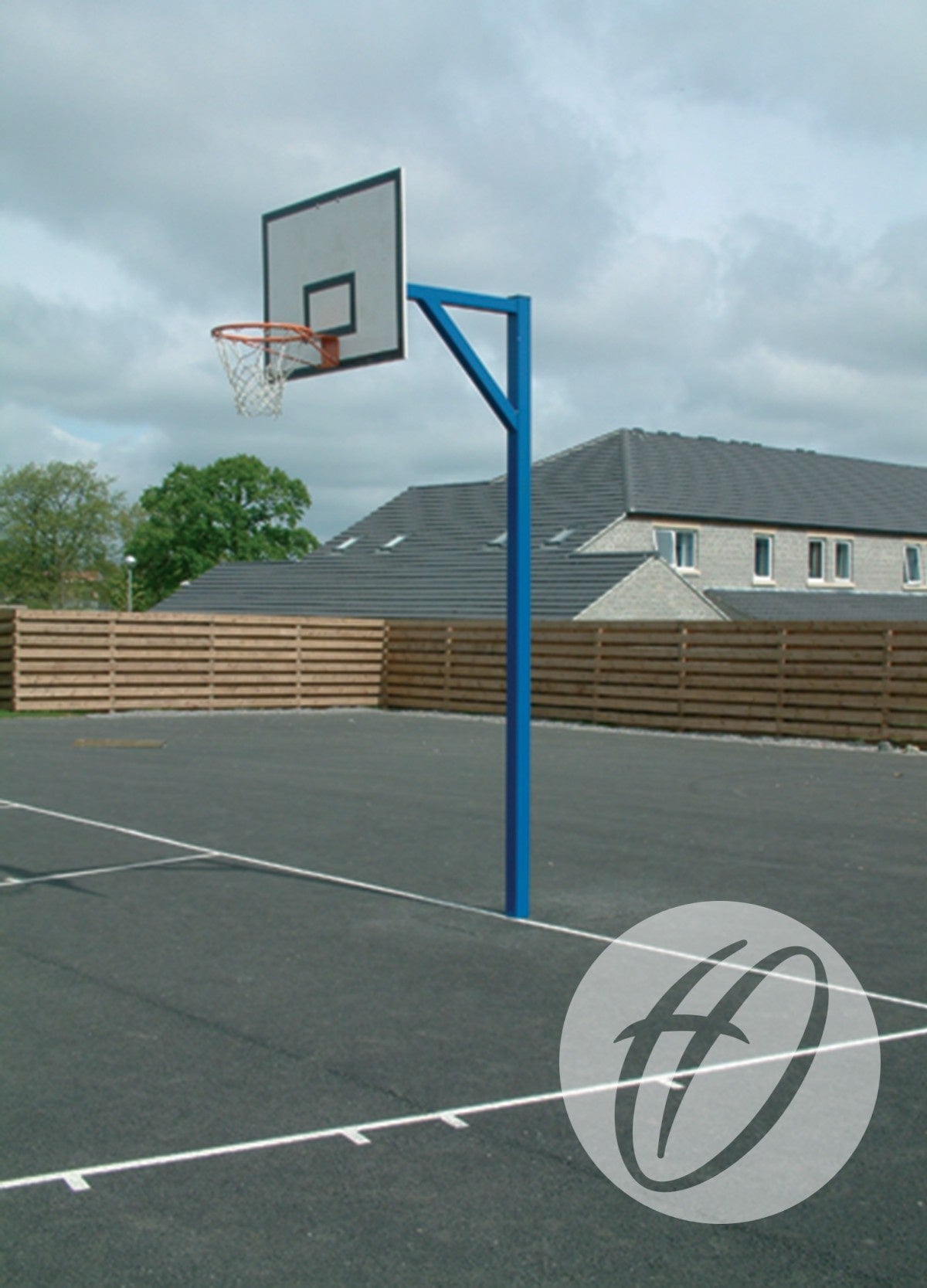 A Harrod Sport Steel Backboards - Heavy Duty Goals blue outdoor basketball hoop stands on a paved court, bordered by a wooden fence and residential homes beneath a cloudy sky.