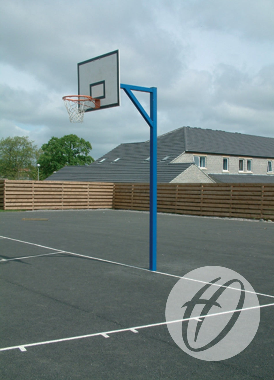 A Harrod Sport Steel Backboards - Heavy Duty Goals blue outdoor basketball hoop stands on a paved court, bordered by a wooden fence and residential homes beneath a cloudy sky.