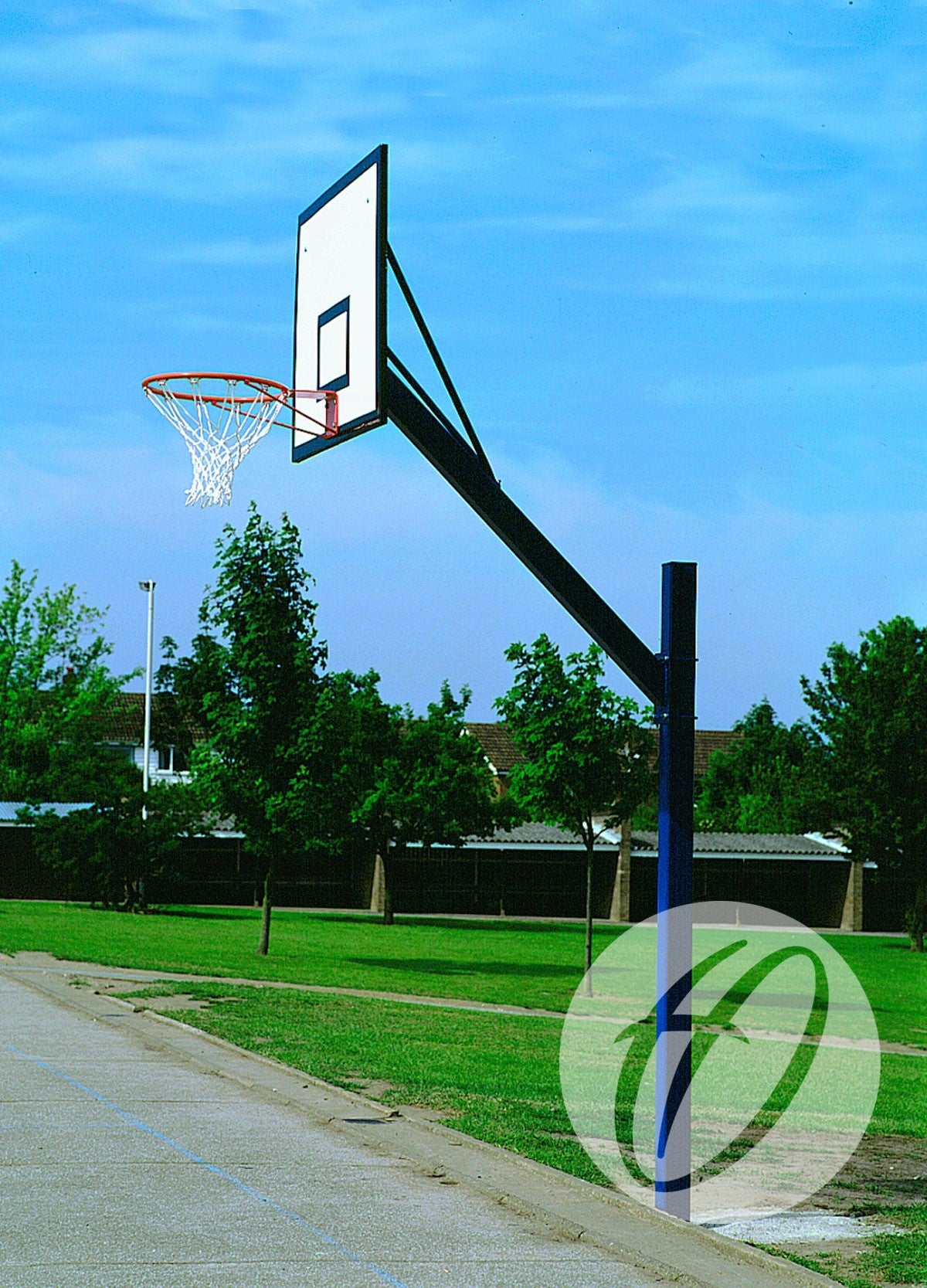 The Harrod Sport Cantilever Basketball Goals with a steel backboard stand at the edge of an outdoor court, surrounded by grass, trees, and a blue sky.