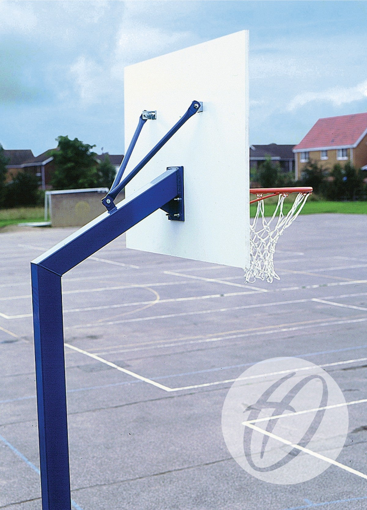 A Harrod Sport Mini Basketball Goals - Permanent with a white backboard stands on an empty court. The net hangs loosely, and houses are visible in the background beneath a partly cloudy sky.