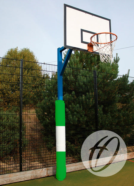 A basketball hoop with a white backboard and rusted rim stands outdoors by a tall black fence and trees. The pole is fitted with Harrod Sport Basketball Post Protectors - Single Coloured in green and white for enhanced safety.