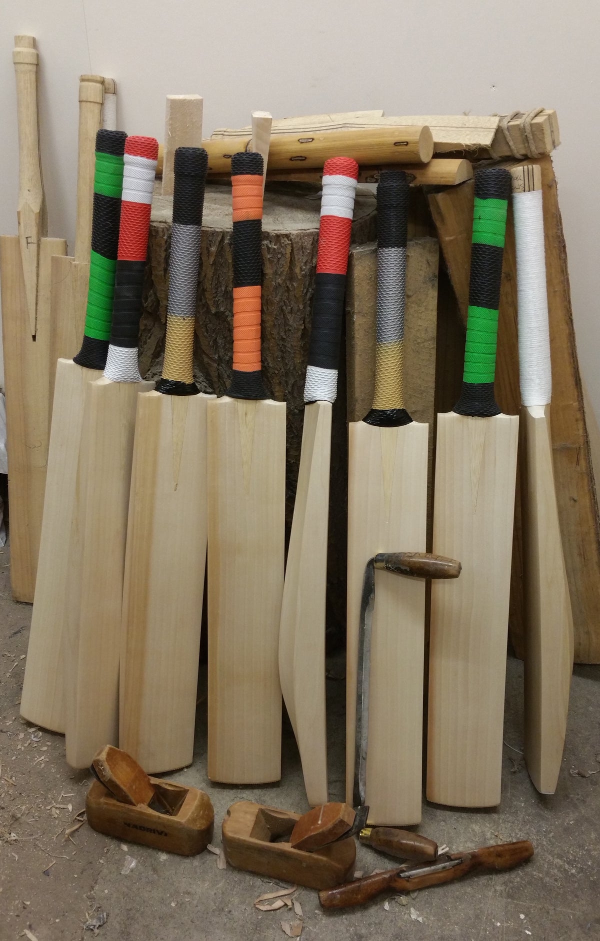 Six wooden cricket bats with colorful handles stand upright by a large tree stump, surrounded by wood shavings and Martin Berrill Sports Cricket Bat Weight Reduction or Rebalance tools used for professional assessment.