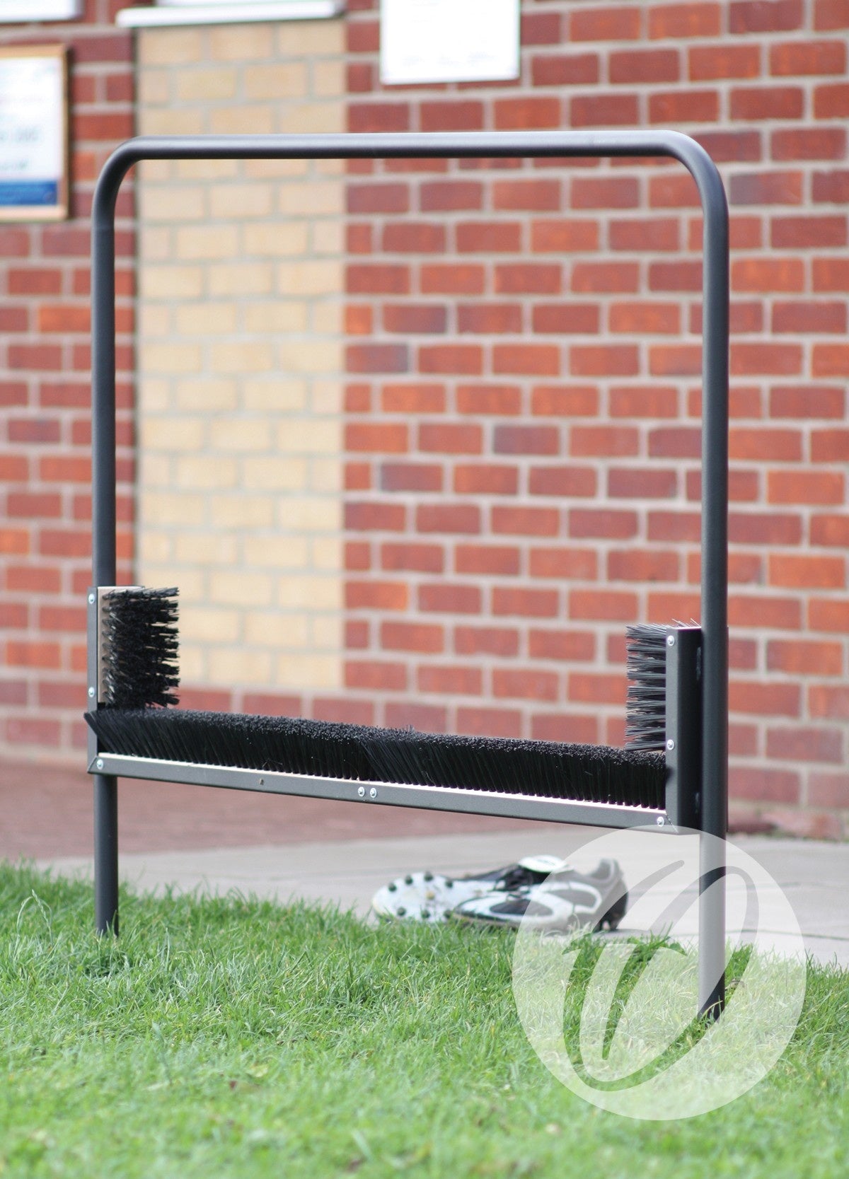 The Harrod Sport Multi Boot Wiper features horizontal and vertical bristles to clean sports shoes. It stands on grass in front of a brick wall, scrubbing off dirt while a pair of shoes rests behind the sturdy steel frame.