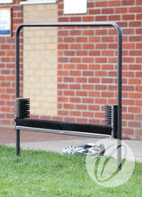 The Harrod Sport Multi Boot Wiper features horizontal and vertical bristles to clean sports shoes. It stands on grass in front of a brick wall, scrubbing off dirt while a pair of shoes rests behind the sturdy steel frame.