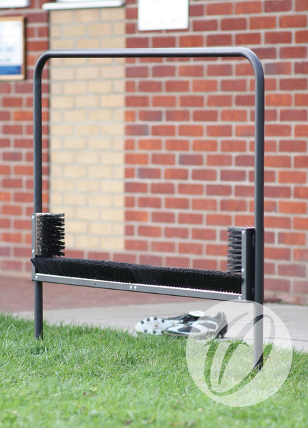 The Harrod Sport Multi Boot Wiper features horizontal and vertical bristles to clean sports shoes. It stands on grass in front of a brick wall, scrubbing off dirt while a pair of shoes rests behind the sturdy steel frame.