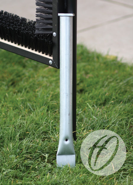 A close-up of a metal Socket For Multi Boot Wipers by Harrod Sport, supporting black-bristled units on green grass near pavement. A transparent circular logo appears in the lower right corner.
