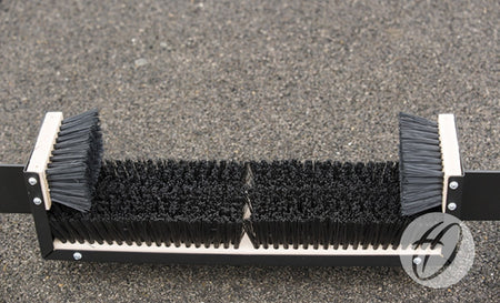 A close-up of the Harrod Sport Fixed Premier Boot Wiper 1.1m, featuring stiff black bristles mounted on a durable rectangular frame and positioned on a rough outdoor surface—perfect for placement near communal changing areas.
