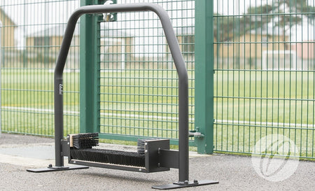A Harrod Sport Freestanding Premier Boot Wiper 1.1m with sturdy boot brushes stands on pavement in front of a green metal fence, with a sports field and buildings visible in the background.