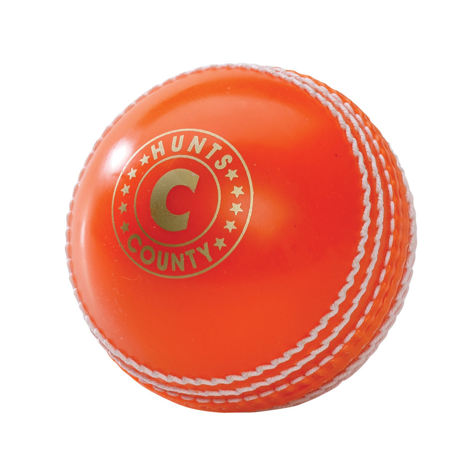 The Hunts County Bomber Ball is an orange cricket training ball from Hunts County, featuring white stitching and a gold logo, displayed on a plain white background.