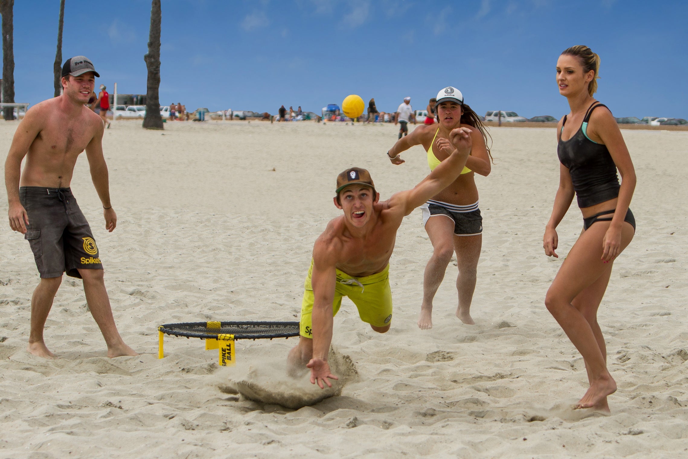 Four young adults play Kankakee Spikeball’s Spikeball Standard Set on a sandy beach. One man dives for the ball near the adjustable net while the others respond, all under a bright, clear blue sky.