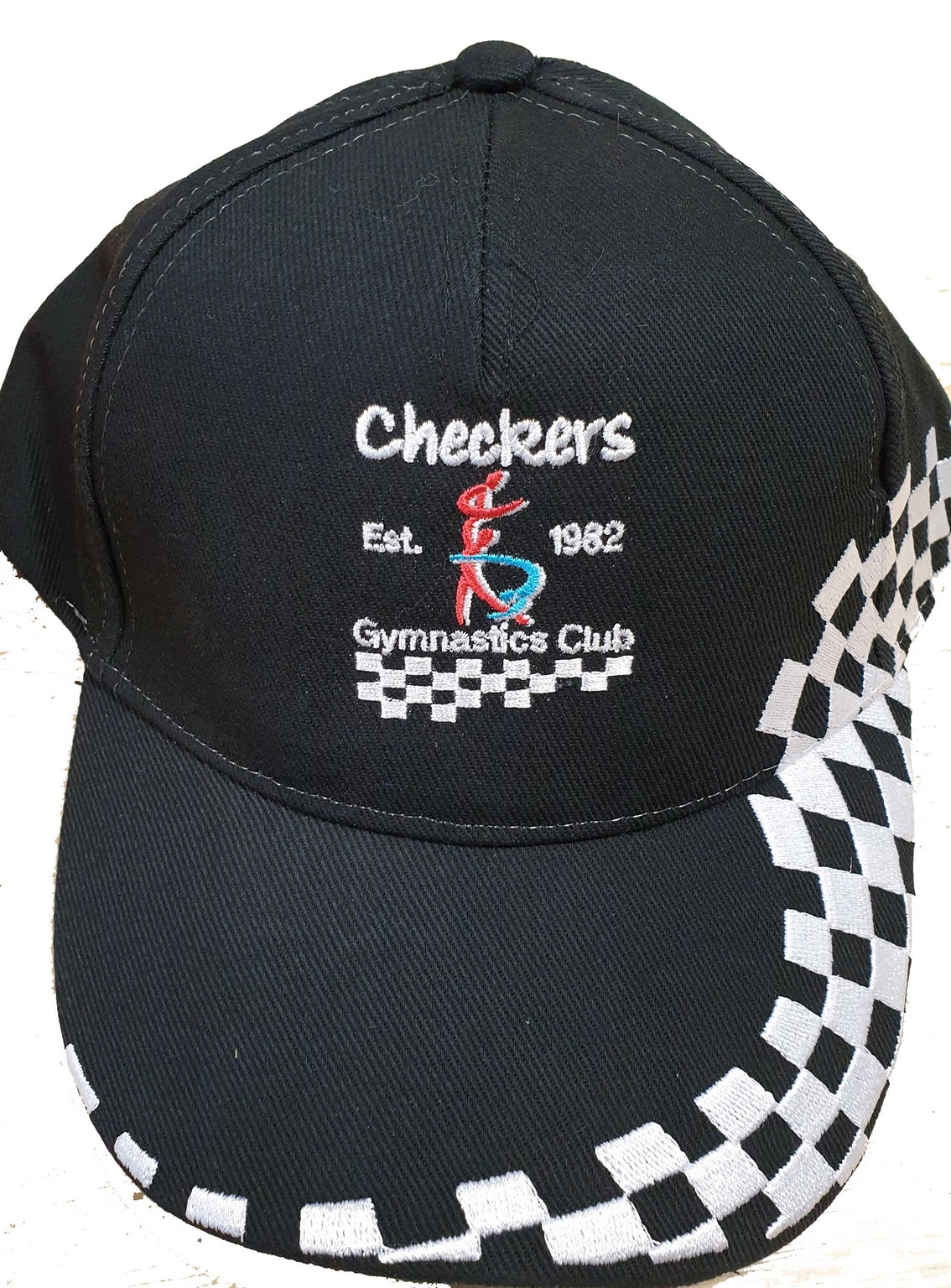 The Checkers Gymnastics Cap by Martin Berrill Sports is a black adjustable cap featuring “Checkers Est. 1982 Gymnastics Club” embroidery, a colorful gymnast figure, and a white checkered pattern on the brim and side.