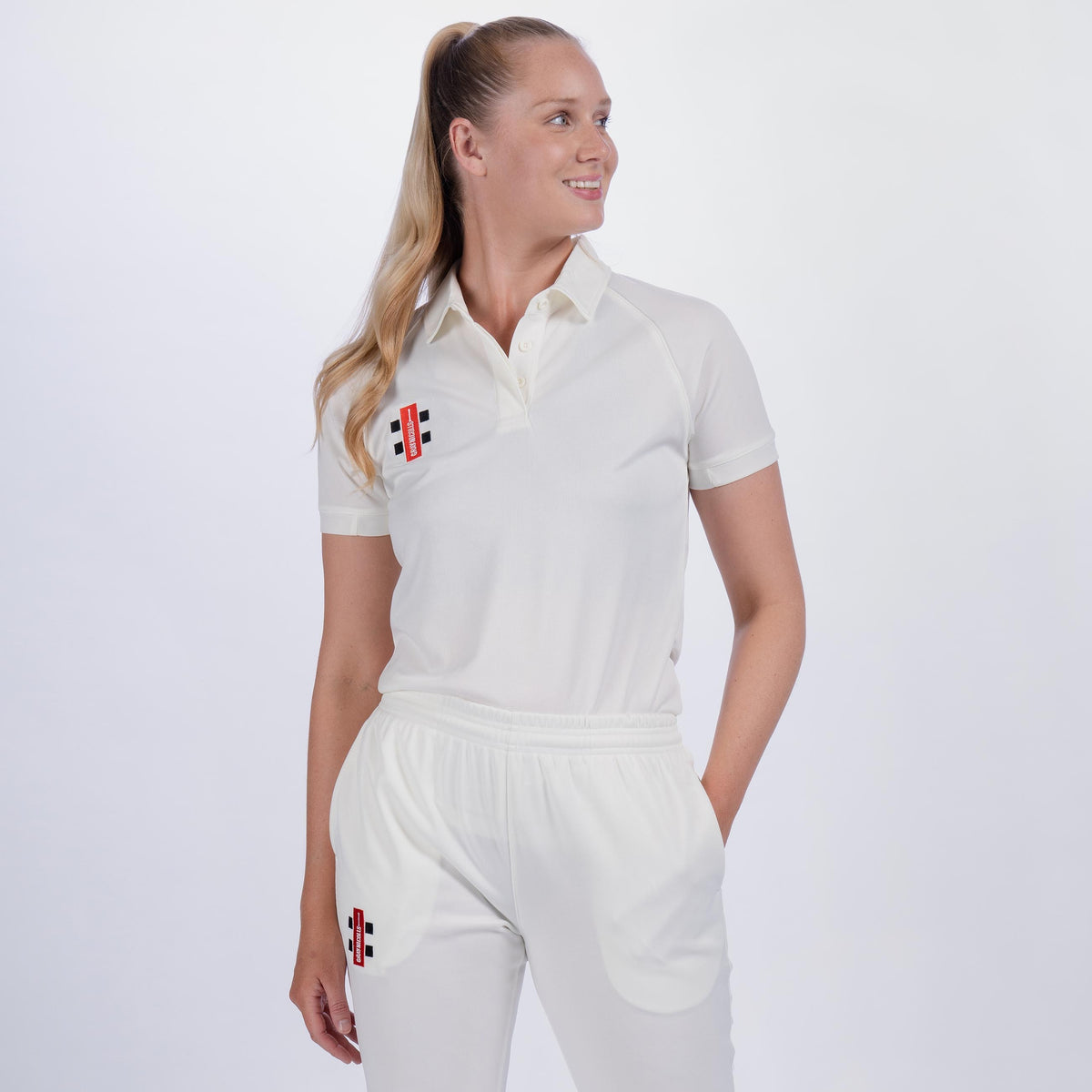 A woman with long blonde hair in a ponytail wears the Gray Nicolls Ladies Matrix V2 Match Shirt, featuring breathable, moisture-wicking fabric and black and red logos. She stands smiling to the side with one hand in her pocket against a plain background.
