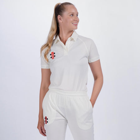 A woman with long blonde hair in a ponytail wears the Gray Nicolls Ladies Matrix V2 Match Shirt, featuring breathable, moisture-wicking fabric and black and red logos. She stands smiling to the side with one hand in her pocket against a plain background.