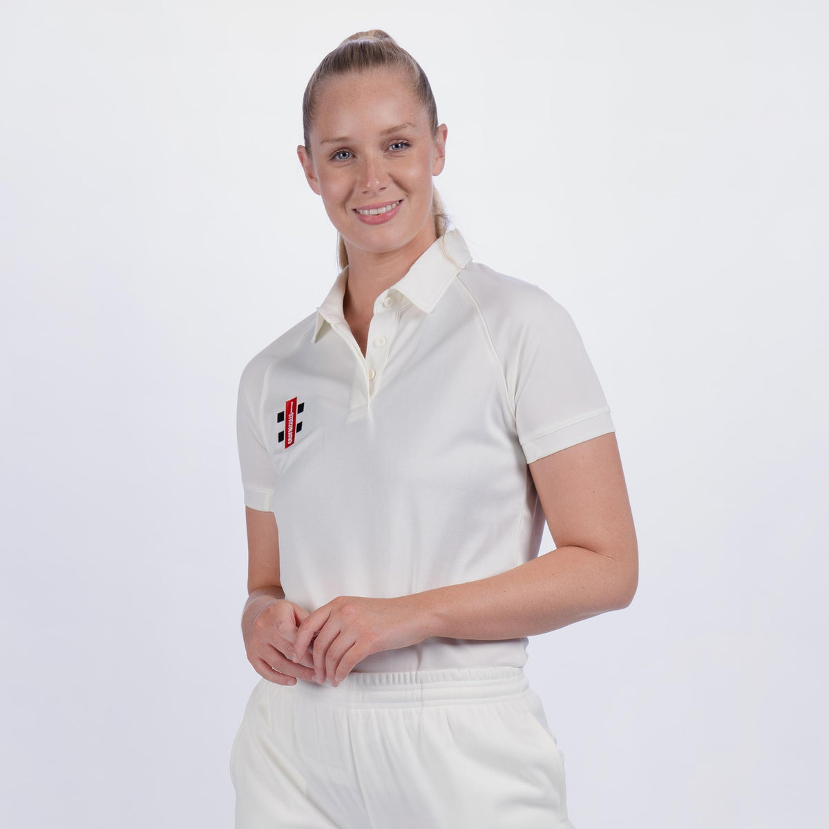 A woman smiles with hands clasped, wearing a Gray Nicolls Ladies Matrix V2 Match Shirt—white, short-sleeved, breathable, with a black, red, and white left-chest emblem. She also wears matching pants against a plain white background.