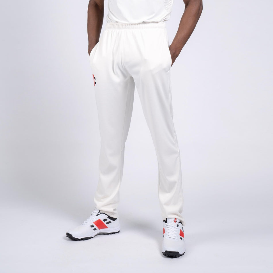 A person stands with hands in pockets, wearing Gray Nicolls Rockhampton CC Pro Performance Cricket Trouser and white sneakers with red and black accents, set against a plain white background. The upper body is partially out of frame.