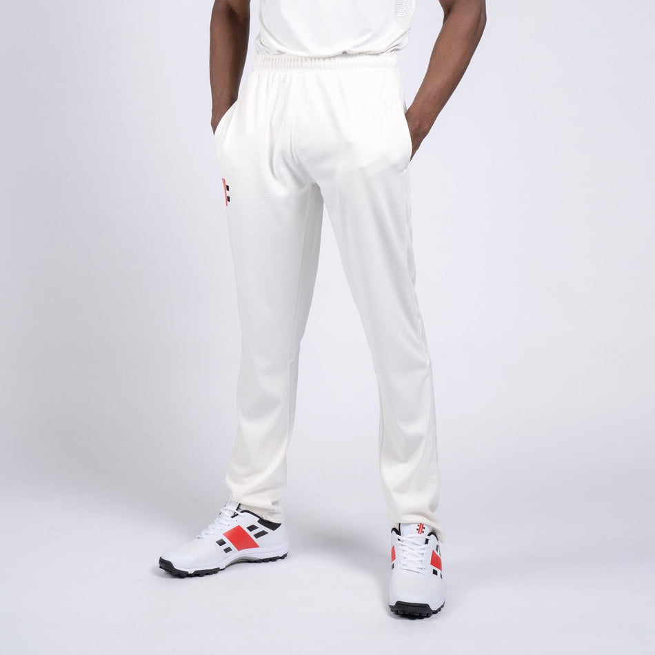 A person wears Gray Nicolls Penallt & Redbrook CC Pro Performance Cricket Trousers with white sneakers featuring red and black accents, standing hands-in-pockets against a white background. Moisture-wicking fabric suits club players.