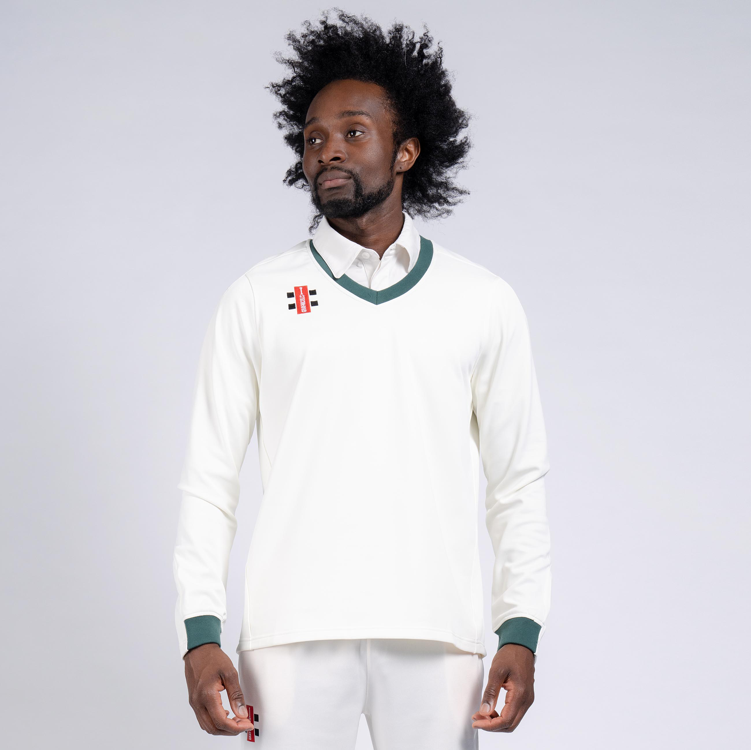 A man with an afro hairstyle wears the Gray Nicolls Oldlands CC Pro Performance Sweater, featuring green trim and a small club logo, over a collared shirt, standing against a plain light background.