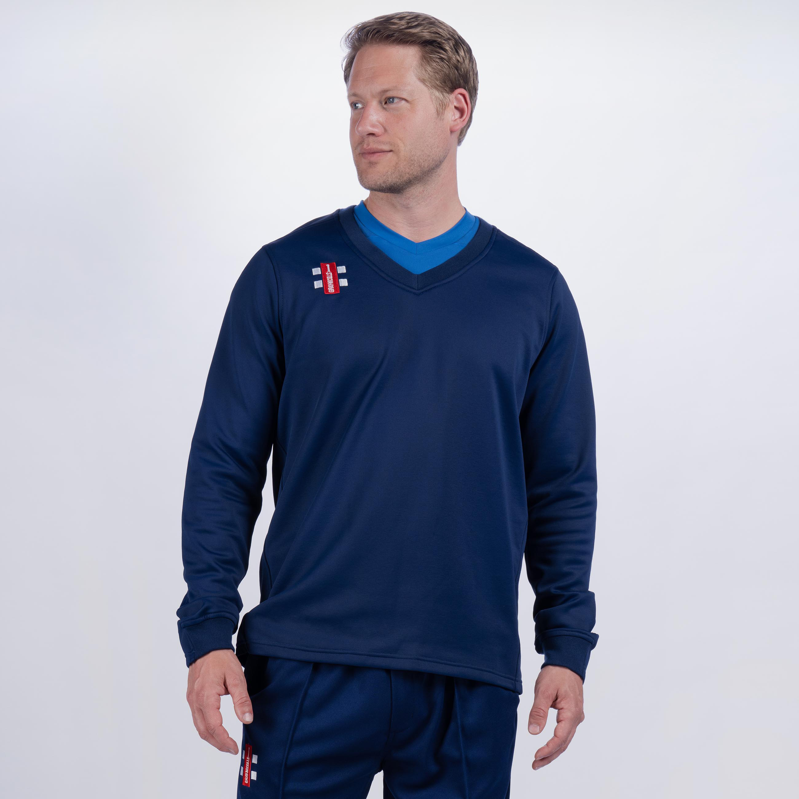 A man wearing a navy blue Gray Nicolls Monmouth CC Pro Performance Sweater and matching pants with small red and white logos stands against a plain light background, looking to his left with a neutral expression.