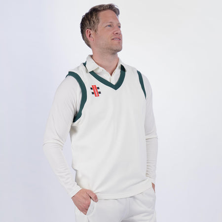 A man wears a Gray Nicolls Oldlands CC Pro Performance Slipover over a white cricket uniform, hands in pockets, looking upward and smiling against a plain light background.